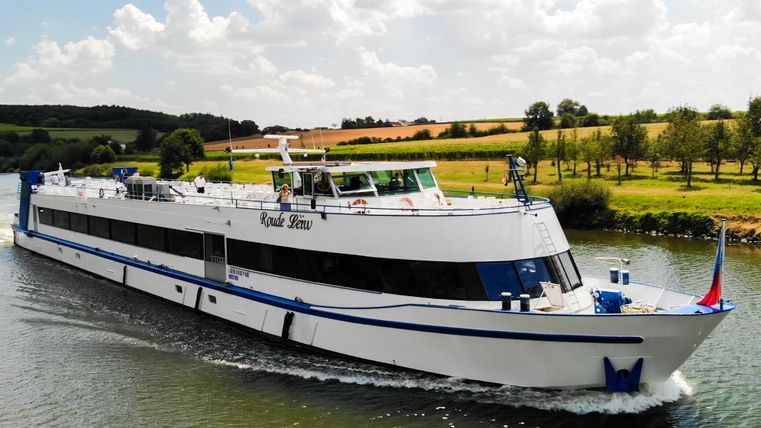Een wit schip vaart over een rustige rivier. Op de achtergrond zijn groene velden en bomen te zien.