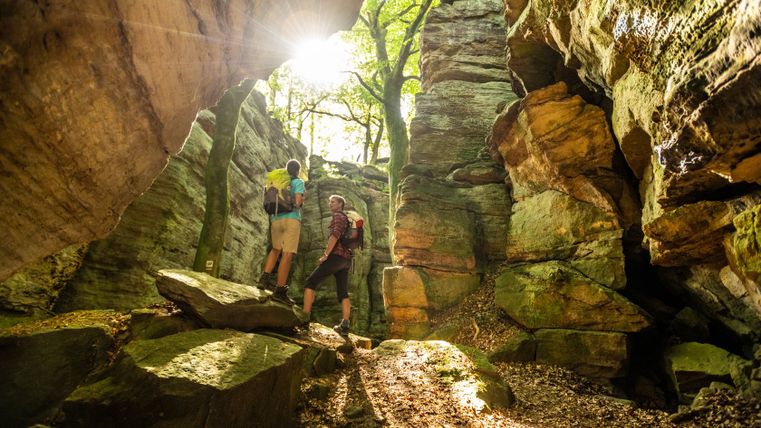 Zwei Wanderer in einer felsigen Schlucht mit Sonnenlicht und Bäumen.