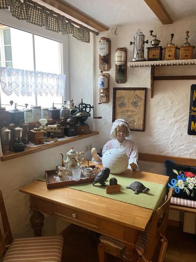 A cozy room with a table and various decorative items. A doll in traditional clothing sits in the corner, giving the room a nostalgic charm.