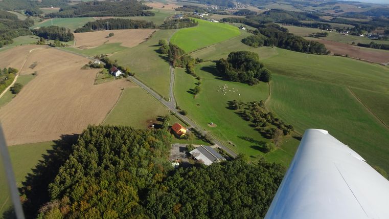 An aerial shot of a rural landscape with fields and forests. The image shows some buildings and a road winding through the green surroundings.