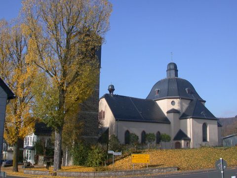 Une église avec un toit rond et une haute tour, entourée d'arbres en automne. Les couleurs des feuilles sont dorées et le ciel est bleu clair.