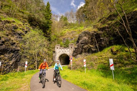 Zwei Radfahrer genießen eine malerische Landschaft auf einem Radweg. Im Hintergrund ist ein alter Tunnel und grüne Hügel zu sehen.