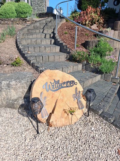 Une pancarte en bois portant l'inscription "Bienvenue" se tient devant un escalier. En arrière-plan, on peut voir des plantes et des pierres.