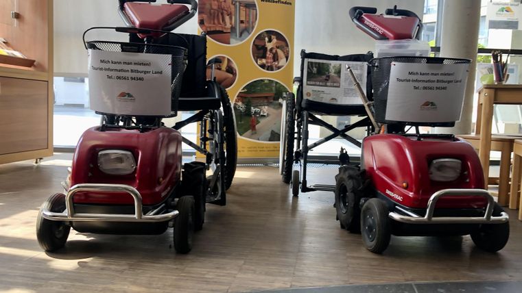Two red motorized wheelchairs are standing next to each other in an interior space. In the background, informational posters can be seen.