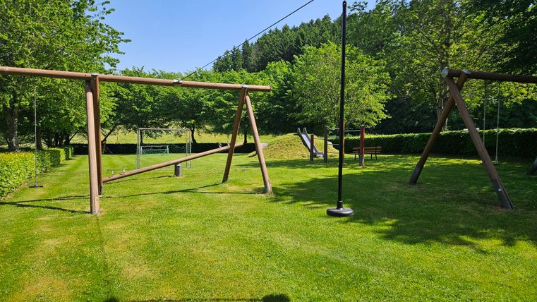 Un terrain de jeu avec des balançoires se trouve sur une pelouse verte. Des arbres entourent la zone et le ciel est d'un bleu clair.