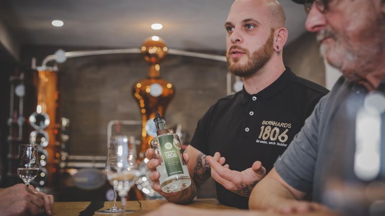 A young man is explaining something at a bar with a drink in his hand. In the background, distillation equipment is visible.