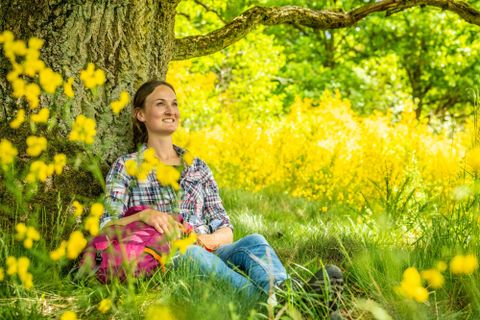 Eine Frau sitzt lächelnd unter einem Baum und genießt die Natur. Rundum sind gelbe Blumen und grünes Gras.