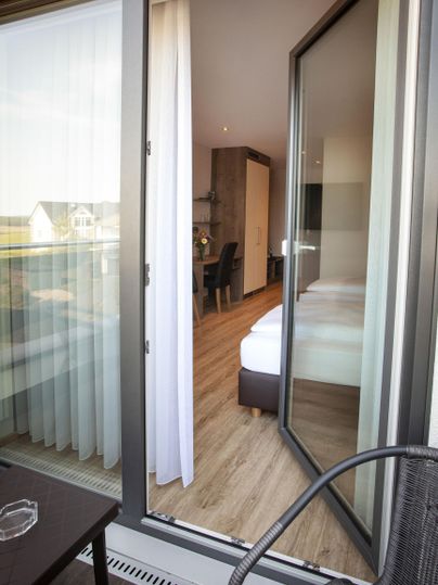 A view of a balcony overlooking a bright room. The doors are wide open and the floor is made of wood.