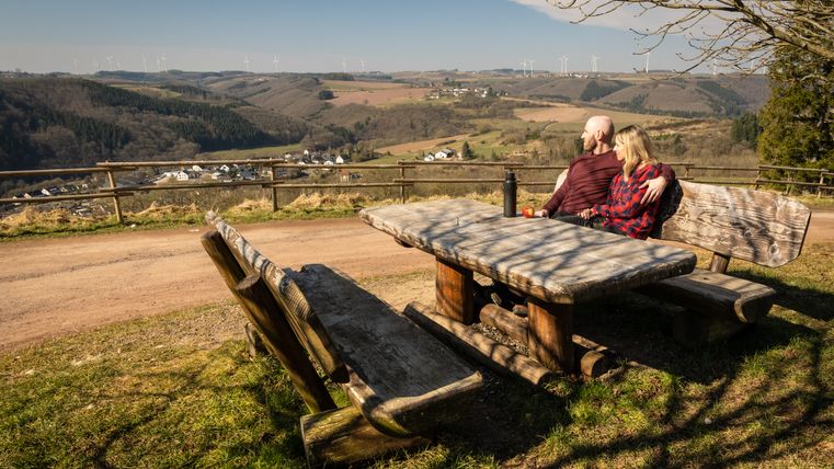 A couple sits on a bench at a wooden table with a view of a hilly landscape and wind turbines in the distance.