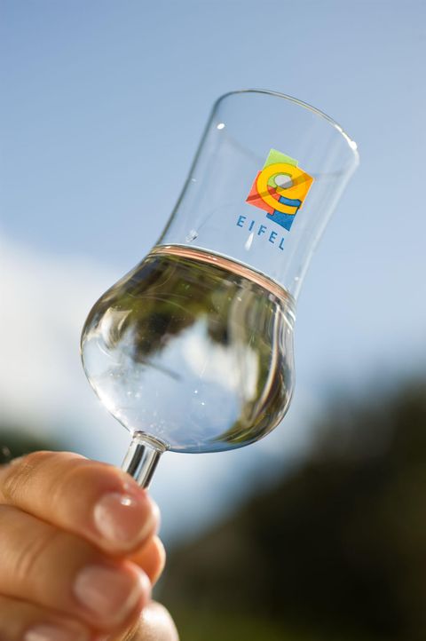 A transparent glass with water is held by a hand. The glass features a colorful logo with the name "Eifel".