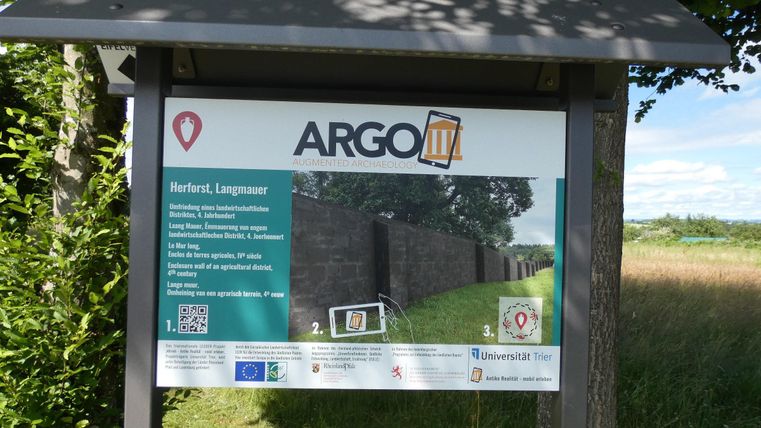 A sign with information about the ARGO III project in Herford. In the background, a historical wall can be seen, surrounded by grass and trees.