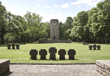 Ein Kriegsdenkmal mit Steinkreuzen auf einer Wiese, ein Turm im Hintergrund, umgeben von gr&uuml;nen B&auml;umen unter blauem Himmel., &copy; TI Bitburger Land