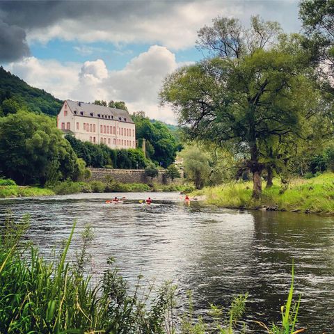 A tranquil river with kayakers and lush trees along the bank. In the background, a historic building stands beneath a cloudy sky.