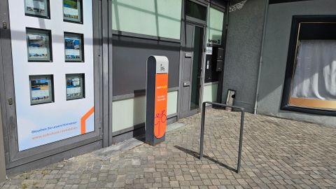 Un parking moderne avec un support à vélos et un stand d'information. La façade du bâtiment présente des fenêtres publicitaires avec des images.