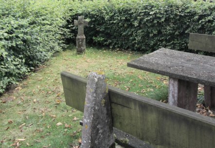 Een stenen kruis staat op een weiland naast een verweerde houten tafel, omringd door dichte heggen en bomen., &copy; M. Bach
