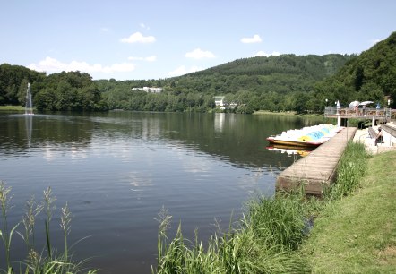 Le lac de barrage de Bitburg aux eaux calmes, entour&eacute; de collines verdoyantes. Des p&eacute;dalos sont amarr&eacute;s au ponton, des gens se d&eacute;tendent sur la rive. Une belle journ&eacute;e d'&eacute;t&eacute;., &copy; TI Bitburger Land