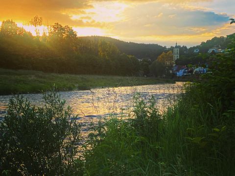 A calm river flows through a green landscape. In the background, the sun shines behind gentle hills and buildings.