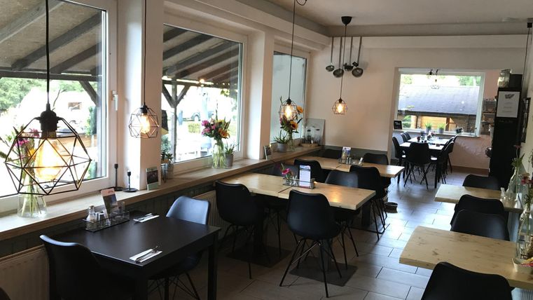 Een modern restaurant met gezellige tafels en stoelen. Grote ramen zorgen voor veel daglicht en een uitnodigende sfeer.