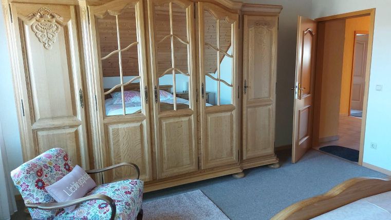 A cozy bedroom with a large wooden wardrobe and a blooming chair. The walls are painted light, and there is access to another room.