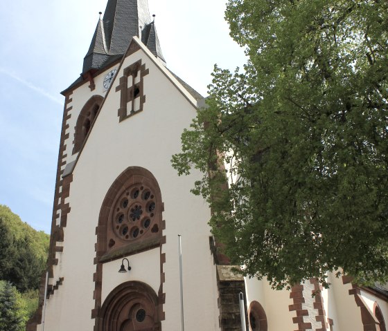 Die Kirche St. Quirinus in Malberg mit ihrem markanten spitzen Turm und einem gro&szlig;en gr&uuml;nen Baum im Vordergrund., &copy; TI Bitburger Land