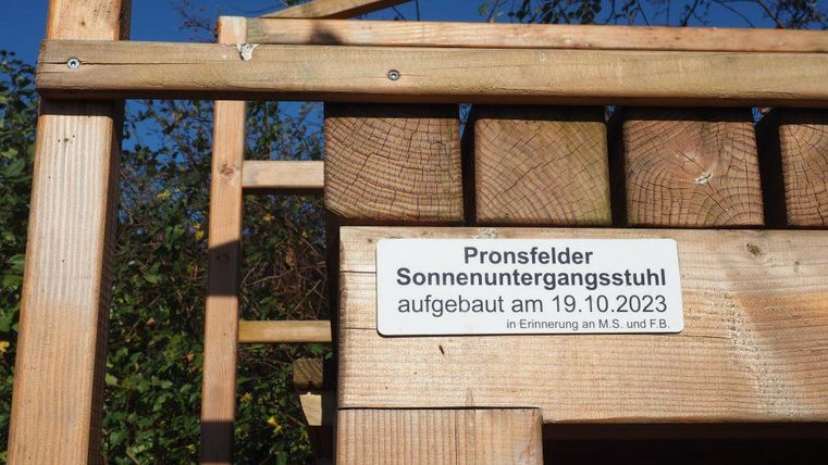A wooden chair with the inscription "Pronsfelder Sunset Chair," which was set up on October 19, 2023. In the background, there are trees and a blue sky visible.