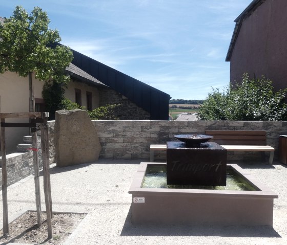 Une petite aire de repos &agrave; Trimport avec une fontaine, un banc en bois et un arbre. En arri&egrave;re-plan, on aper&ccedil;oit des b&acirc;timents et un ciel bleu., &copy; TI Bitburger Land