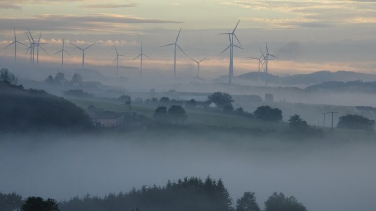 A foggy landscape with wind turbines in the background. The sky is overcast, and the mist gives the scene a mystical atmosphere.