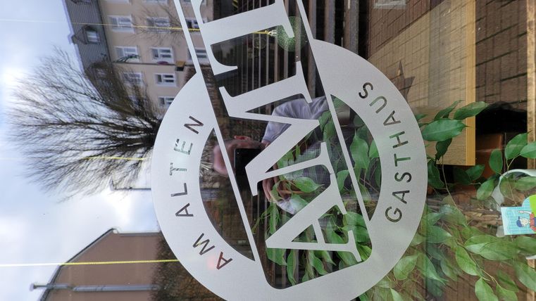 Une fenêtre avec le logo de l'auberge "Am Alten". En arrière-plan, des plantes et une rue sont visibles.
