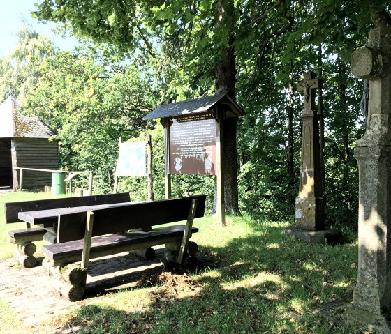 Rastplatz mit Holzb&auml;nken, Infotafel und Steinkreuz unter B&auml;umen. Im Hintergrund eine Holzh&uuml;tte und gr&uuml;ne Vegetation., &copy; Tourist-Info Bitburger Land M.Mayer