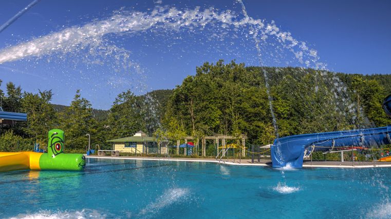 Ein Schwimmbecken mit klar blauem Wasser und Wasserspielen. Im Hintergrund sind grüne Bäume und eine Rutsche zu sehen.