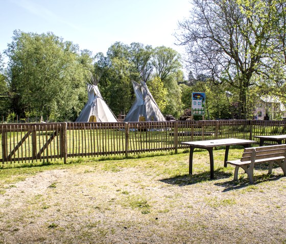 Een speeltuin met twee tipi's achter een houten hek, omringd door bomen. Op de voorgrond staan tafels en banken op een grindvloer., &copy; Tourist-Information Bitburger Land_M. Mayer