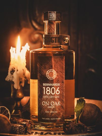 A bottle of Bernhards 1806 Eifel Dry Gin on a wooden tray, surrounded by decorative elements and a burning candle. The warm lighting creates a cozy atmosphere.