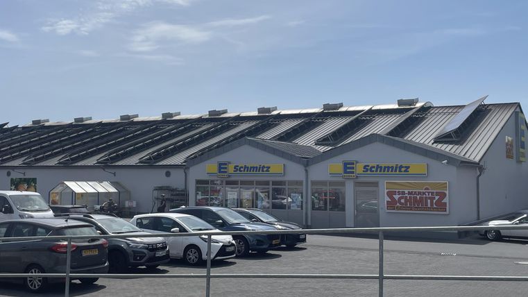 Een kantoorgebouw met de naam "Schmitz" en een modern dak. Voor het gebouw staan meerdere auto’s geparkeerd op een grote parkeerplaats.