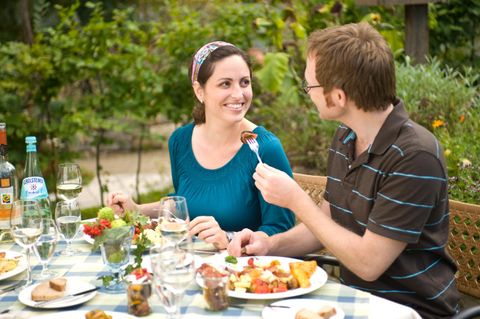 A happy couple enjoying a meal outdoors. The table is set with various dishes and drinks, surrounded by green plants.