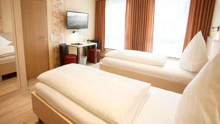 A modern hotel room with two single beds and wooden flooring. There is a television on the wall and a small table with chairs.