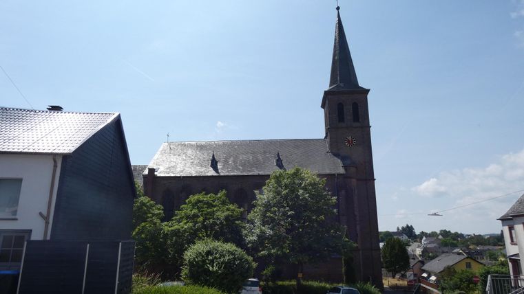 Une vieille église avec un haut clocher se dresse derrière des arbres verts. Le ciel est clair et bleu.
