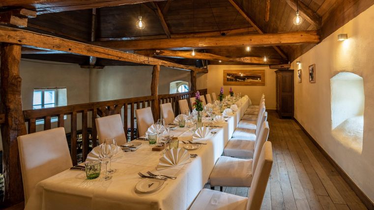 An elegant dining area with long tables and white tablecloths. The wooden beam ceiling and soft lighting create a cozy atmosphere.
