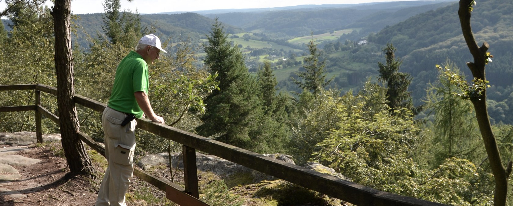 Een man in groene kleding staat bij een reling en kijkt uit over een bebost landschap met heuvels en dalen., &copy; Lauschtour.de