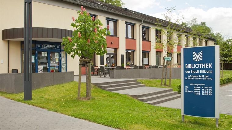 Een modern bibliotheekgebouw met een uitnodigende ingang. Op het terrein zijn bomen en een informatiebord met openingstijden te zien.