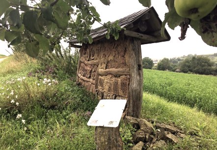 Een insectenhotel van hout en klei staat aan de rand van een groen veld. Een informatiebord geeft informatie over het hotel. Bloeiende planten op de voorgrond., &copy; TI Bitburger Land M.Mayer