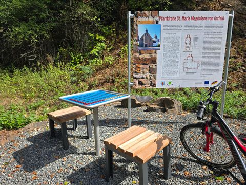 An information kiosk with a sign about St. Mary Magdalene Church stands in the countryside. Next to it are two benches and a bicycle.