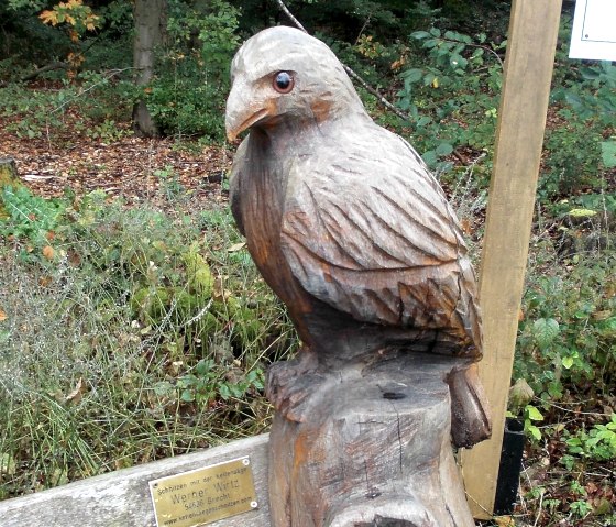 Eine Holzskulptur eines Vogels sitzt auf einem Baumstamm im Wald. Eine Plakette ist an der Skulptur angebracht., &copy; Tourist-Information Bitburger Land