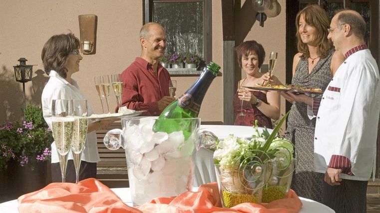 A cheerful group of people is celebrating outdoors with champagne glasses and a beautiful selection of snacks. The table is decorated with a bottle of champagne and ice.