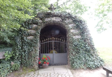 Une grotte de Lourdes recouverte de lierre avec une porte. Des fleurs sont visibles sur le portail. Des panneaux avec des inscriptions d&eacute;corent l'entr&eacute;e., &copy; Eifelverein Ortsgruppe Speicher