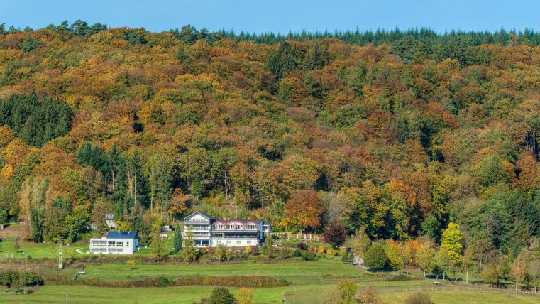 Une vallée pittoresque avec des arbres colorés en automne. Au premier plan se trouvent deux bâtiments, entourés par la nature.