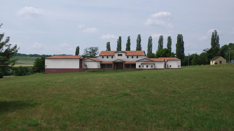 A large, modern building stands on a grassy area. In the background, there are tall trees and a clear sky.