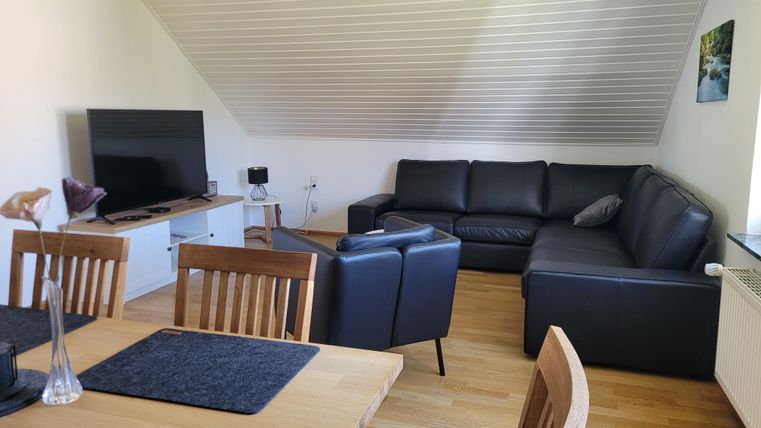 A modern living room with a large black sofa and a flat-screen TV. The room has a wooden-paneled floor and a bright, welcoming atmosphere.