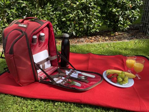 Ein roter Picknickrucksack steht auf einer Decke im Grünen. Darin sind Besteck, Flaschen und ein Bereich mit frischem Obst und Getränken.