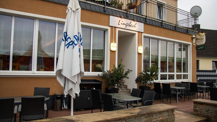 Ein gemütliches Restaurant mit einer Terrasse und mehreren Tischen. Die Fassade ist hell erleuchtet, und ein Sonnenschirm steht vor dem Eingang.