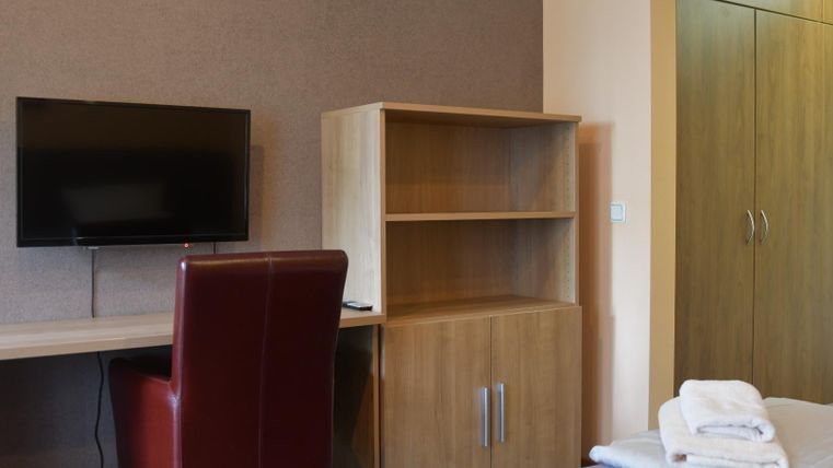 A modern bedroom with a desk, a chair, and a television. In the background, there is a wardrobe and a bed with fresh towels.